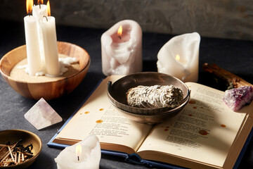 occult science and supernatural concept - magic book, white sage, burning candles and different...