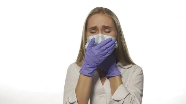 Young nurse in medical mask on white background in robe looks directly into camera. With one hand holds his throat and coughs. She is sick and sad. High quality 4k footage