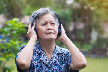 Senior Asian woman wearing wireless headphones listening to a favorite song while standing in a garden. Concept of aged people and healthcare
