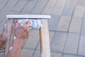 Senior man holding a brush for paint white on an old wooden chair without a backrest. Old chairs...