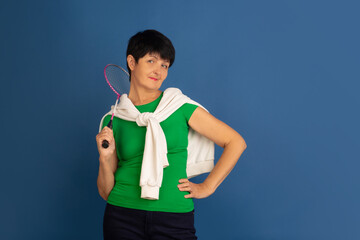 Playing badminton. Portrait of senior woman in stylish outfit isolated on blue studio background....