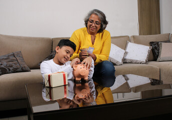 Indian/asian Grandma teaching her grand son importance of savings, putting coins into pink piggy...