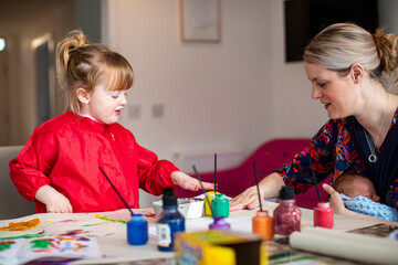 Young girl using paints at kitchen table with mother sitting nearby holding baby