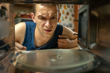 Dark-haired man has opened the microwave oven and looks at it with a surprised look, a photo from inside.