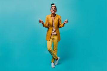 Fototapeta na wymiar Positive african woman in trendy yellow costume smiling to camera. Indoor photo of pretty black girl standing on blue background.
