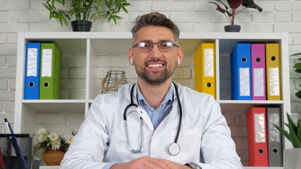 Webcam view smiling man remote doctor wears white medical coat wireless earphones sits on chair at desk in hospital office looks camera consult online distance video call chat laptop computer screen