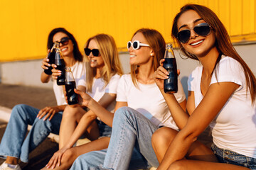 Smiling young women in sunglasses with drinks sit on border against yellow wall in city on sunny summer day