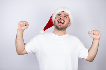 Young handsome man wearing a Santa hat over white background very happy and excited making winner gesture with raised arms, smiling and screaming for success.