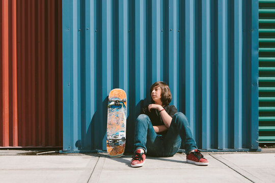 Teenage boy sitting with skateboard against warehouse wall