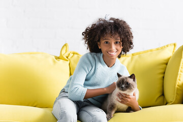 Happy african american girl stroking siamese cat, while sitting on sofa at home