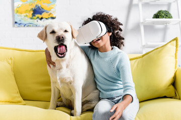 Curly african american girl in vr headset embracing dog yawning, while sitting on sofa on blurred background