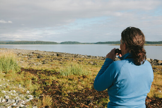 Middle aged woman using binoculars, looking towards remote coastline of an inlet on the Alaska coastline. 