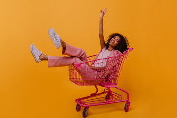 Chilling african girl posing in shopping cart. Indoor photo of emotional young lady in pink clothes. - obrazy, fototapety, plakaty