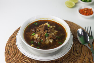 Rawon, Indonesian traditional beef soup from Java Island. Served on white ceramic bowl topped with onion cracker and spring onion,chili paste and lime slices on rattan matt. Isolated white background.