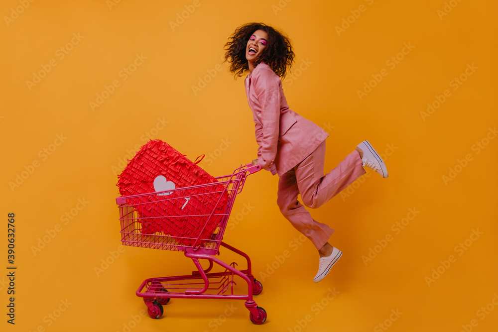 Wall mural Enthusiastic shopaholic girl jumping on yellow background. Trendy african woman posing with shopping cart.