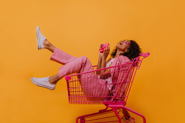 Laughing woman in white gumshoes sitting in shopping cart. Studio photo of amazing black girl with...