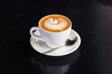 a white cup with cappuccino coffee with beautiful latte art stands on a white plate along with a teaspoon. a cup of coffee stands on a black table