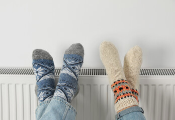 Couple warming legs on heating radiator near white wall, closeup