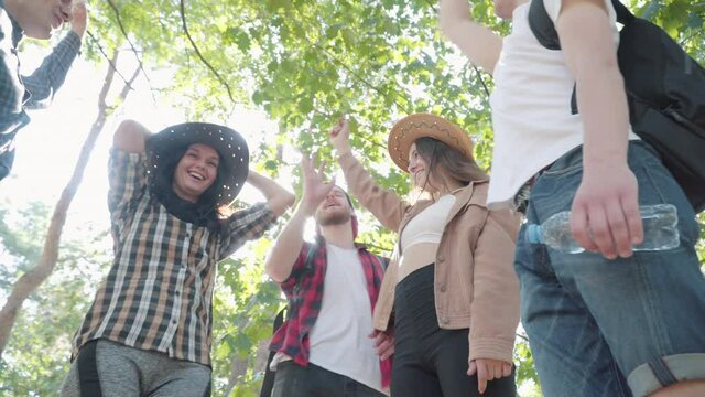 Bottom angle view of positive Caucasian young men and women stacking hands in sunlight outdoors. Handstack of group of happy smiling relaxed tourists on summer day. Joy and tourism concept.