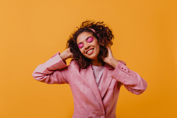 Good-humoured wonderful woman in pink jacket enjoying photoshoot. Debonair young lady with curly hair dancing on orange background.