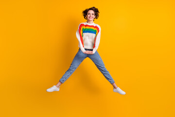 Fototapeta na wymiar Full size photo of cute cheerful young woman jump wear rainbow sweater denim jeans white footwear isolated on bright yellow background