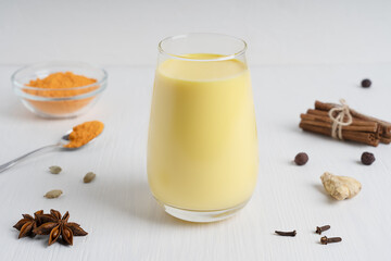 Glass of golden or turmeric milk which is Indian drink used as alternative remedy to boost immunity served with spoon and bowl of curcuma powder, anise and spice on white wooden background. Horizontal