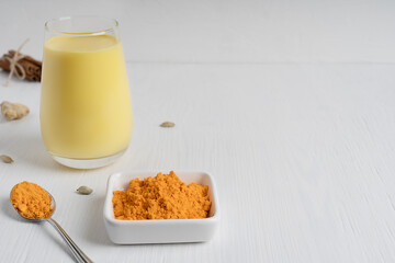Glass of golden or turmeric milk which is homemade drink used as alternative remedy to boost immunity served with spoon and bowl of curcuma powder on white wooden background. Image with copy space