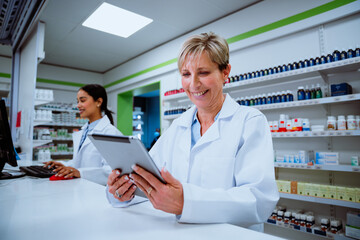 Senior pharmacist scrolling through digital tablet standing behind counter in drugstore waiting for...