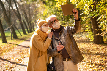 Smiling senior couple in protective mask using digital tablet in autumn park.