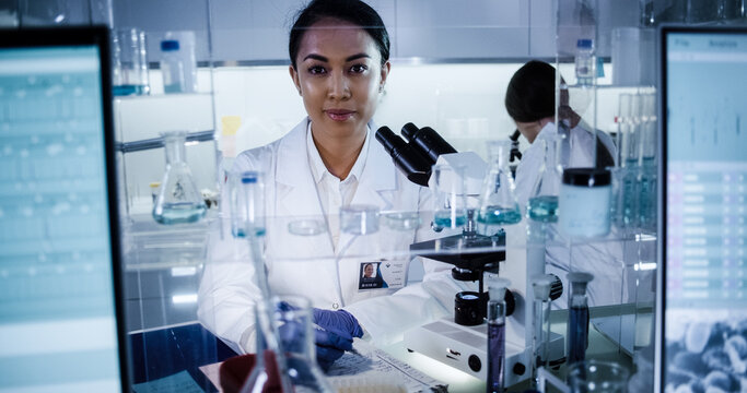 Multi ethnic science team working with microscope and computer. Female doctors in futuristic laboratory. Looking at camera