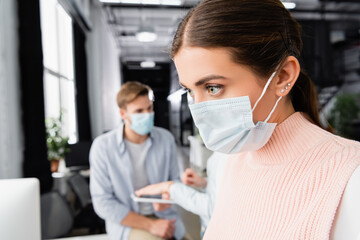 Fototapeta na wymiar Businesswoman in medical mask standing near colleagues on blurred background in office