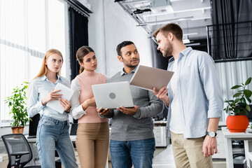 Businessman holding paper folder near multiethnic colleagues with laptop and digital tablet
