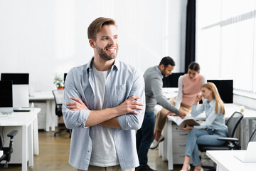 Obraz premium Smiling businessman with crossed arms standing near multicultural colleagues on blurred background in office