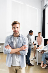Fototapeta premium Young businessman with crossed arms looking at camera near colleagues working on blurred background