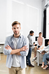 Young businessman with crossed arms looking at camera near colleagues working on blurred background