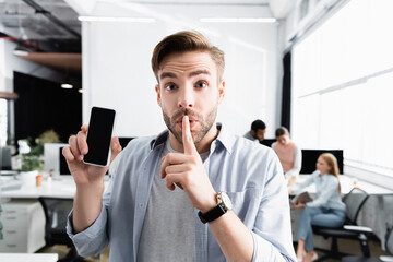 Businessman showing smartphone with blank screen and quiet sign in office