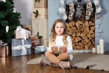 A cute little girl in a white sweater and brown pants sits near the fireplace and dressed Christmas tree and looks into the camera, holding a gift in eco paper