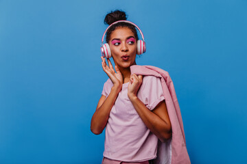 Gorgeous black lady with pink makeup enjoying music on blue background. Indoor photo of carefree girl in casual clothes and headphones.