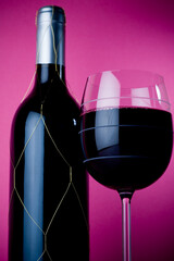Colorful studio shot of black wine bottle and glass filled with dark red wine. Pink background with selective focus.