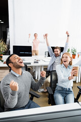 Fototapeta premium Excited indian businessman showing yeah gesture near colleagues and computers on blurred background in office