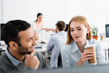Businesswoman with coffee to go sitting near smiling indian businessman and colleagues on blurred background