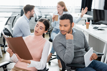 Businesswoman looking at paper folder near indian colleague with digital tablet in office