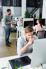 Pensive businessman looking at computer near graphics tablet and multicultural colleagues on blurred background