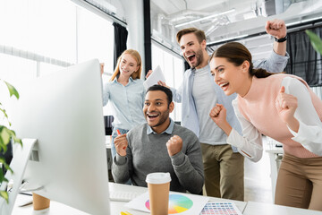 Cheerful multiethnic businesspeople showing yeah gesture near computer, colorful swatches and coffee to go on blurred foreground