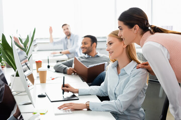 Fototapeta premium Smiling businesswoman using computer and graphics tablet near multiethnic colleagues on blurred foreground in office