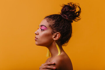 Adorable woman with pink makeup spending time in studio. Serious african lady with yellow earrings posing on orange background.