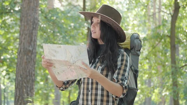 Portrait of beautiful positive Caucasian woman with tourist backpack examining paper map and leaving. Smiling charming female traveler enjoying hiking on sunny summer day outdoors.
