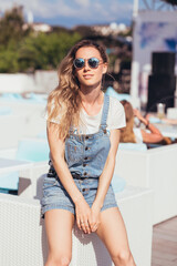 European american young girl with positive emotion sits on lounge in outdoor cafe on sunny summer day. Seaside vacation concept in tropical country. Concept of cheap trip for young people
