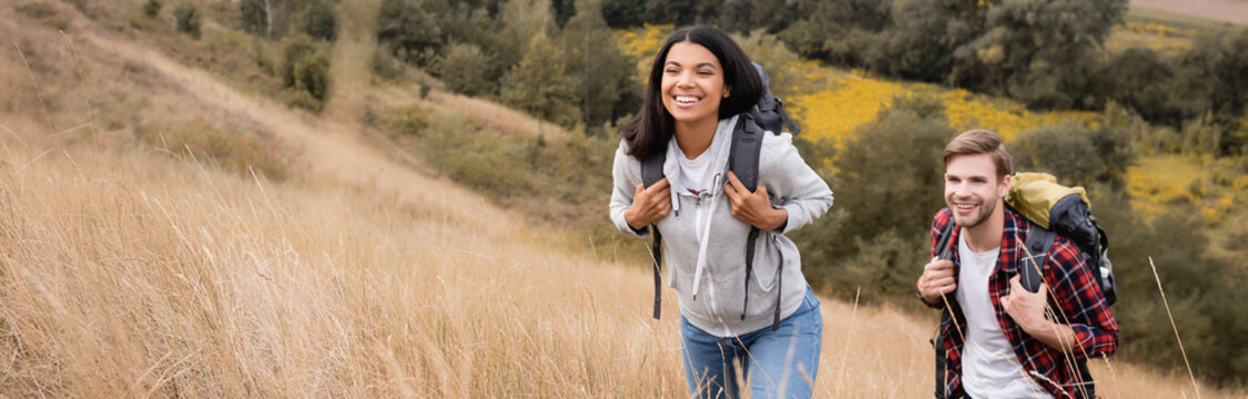 Cheerful multicultural hikers walking with backpacks on field, banner