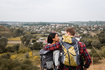 Back view of man embracing african american girlfriend with backpack during trip outdoors